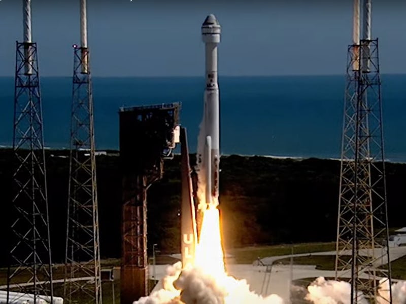 A tall rocket ignites as it lifts off its launch pad. The ocean appears in the background.