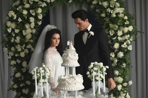 A bride and groom in formal wedding attire stand beside a tiered wedding cake, surrounded by white f...