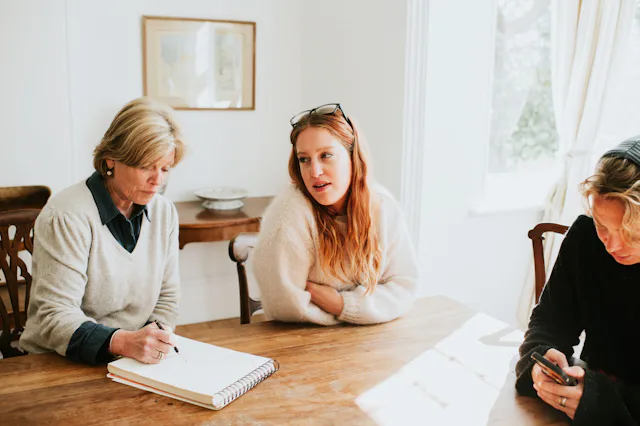 An adult woman sits at a dining table with her mom, who takes notes on a notepad.