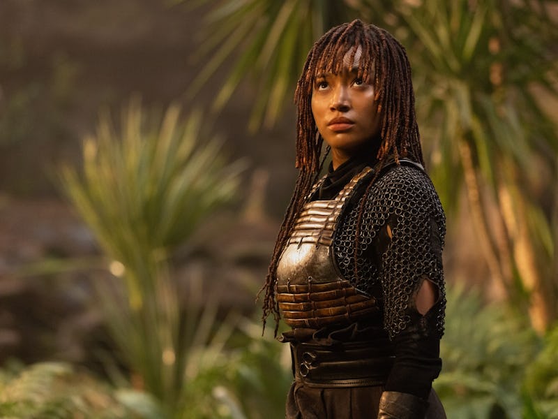 A woman in medieval warrior costume stands in a jungle, looking off into the distance with focus and...