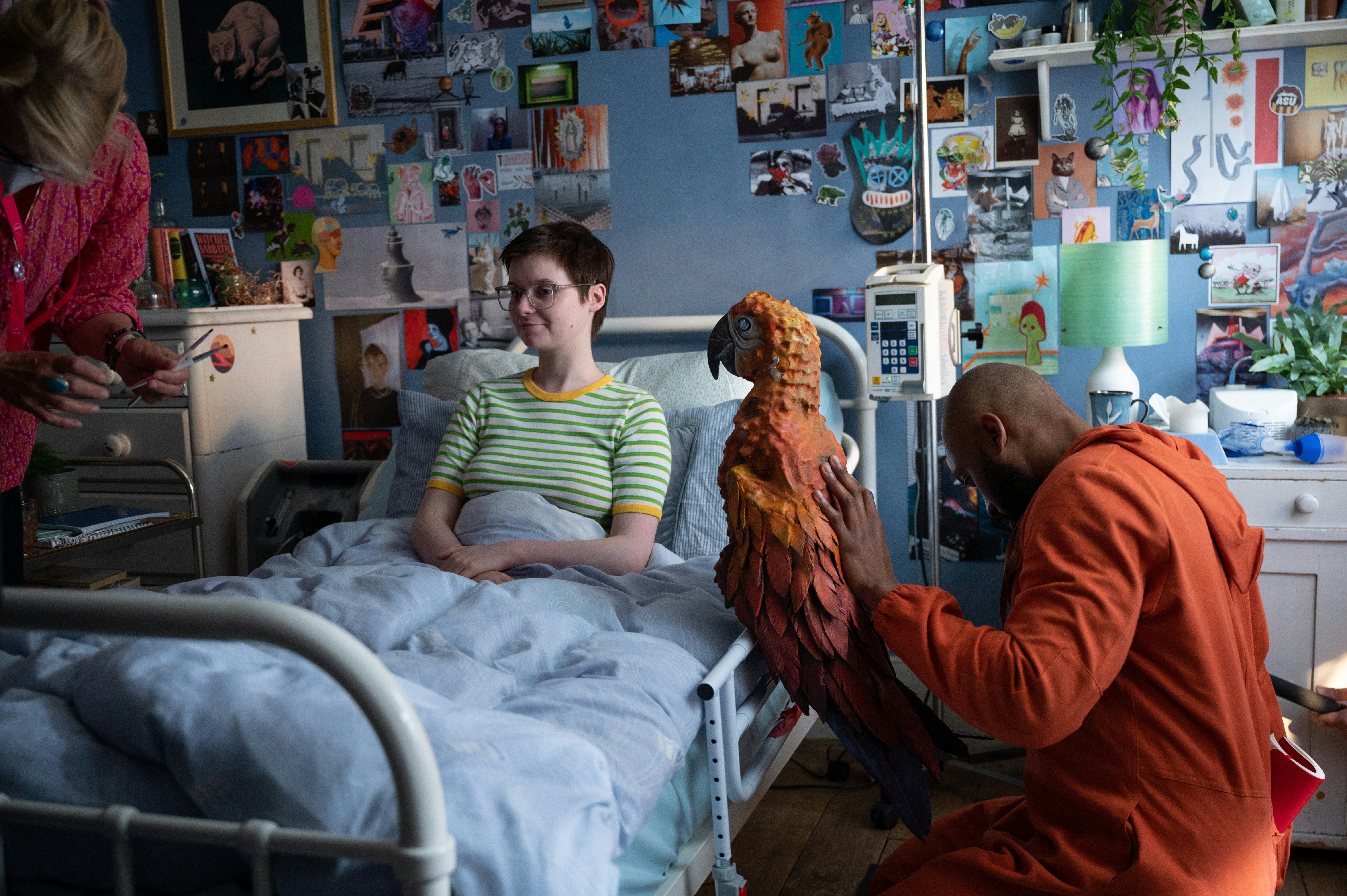 Lola Petticrew as Tuesday in a hospital bed smiles at Arinzé Kene in orange, holding a large parrot&hellip;