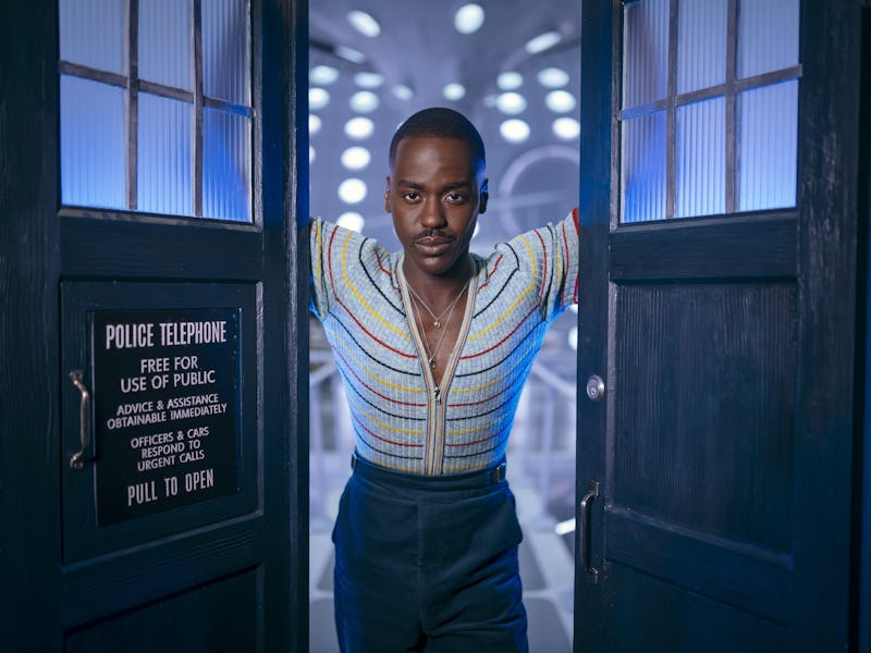 A man stands confidently with arms spread inside a lit, futuristic hallway, framed by blue double do...