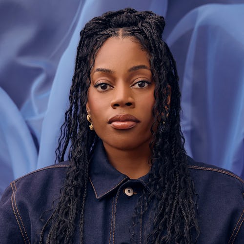 A portrait of a woman with braided hair wearing a dark jacket, against a soft-focus blue fabric back…