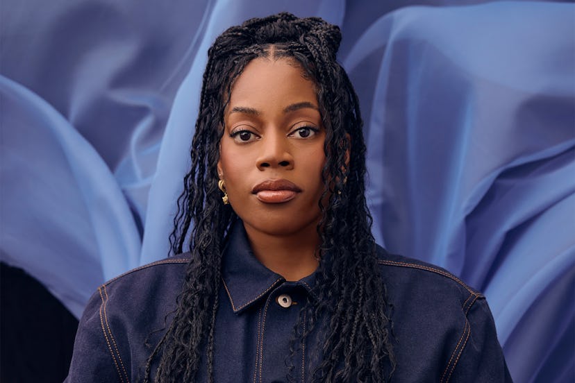A portrait of a woman with braided hair wearing a dark jacket, against a soft-focus blue fabric back...