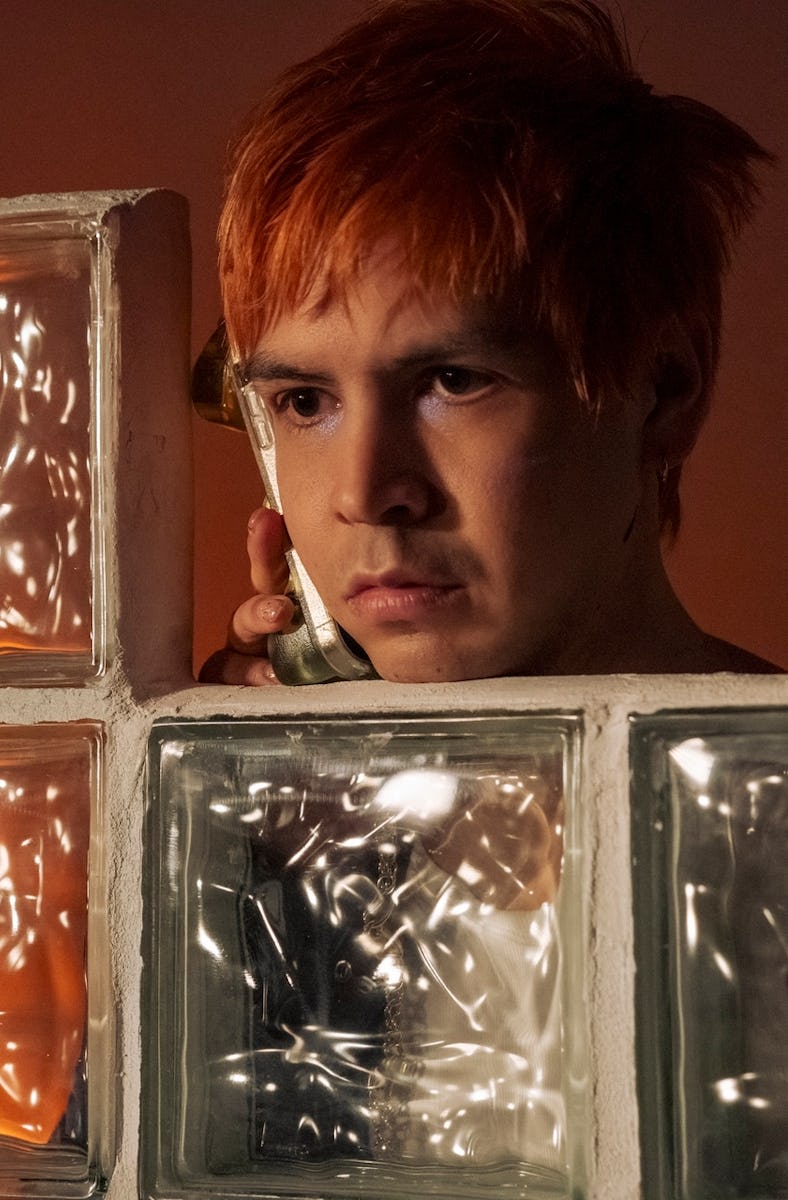 A young man with orange hair peers pensively through a glass block window, with a plant's shadow and...