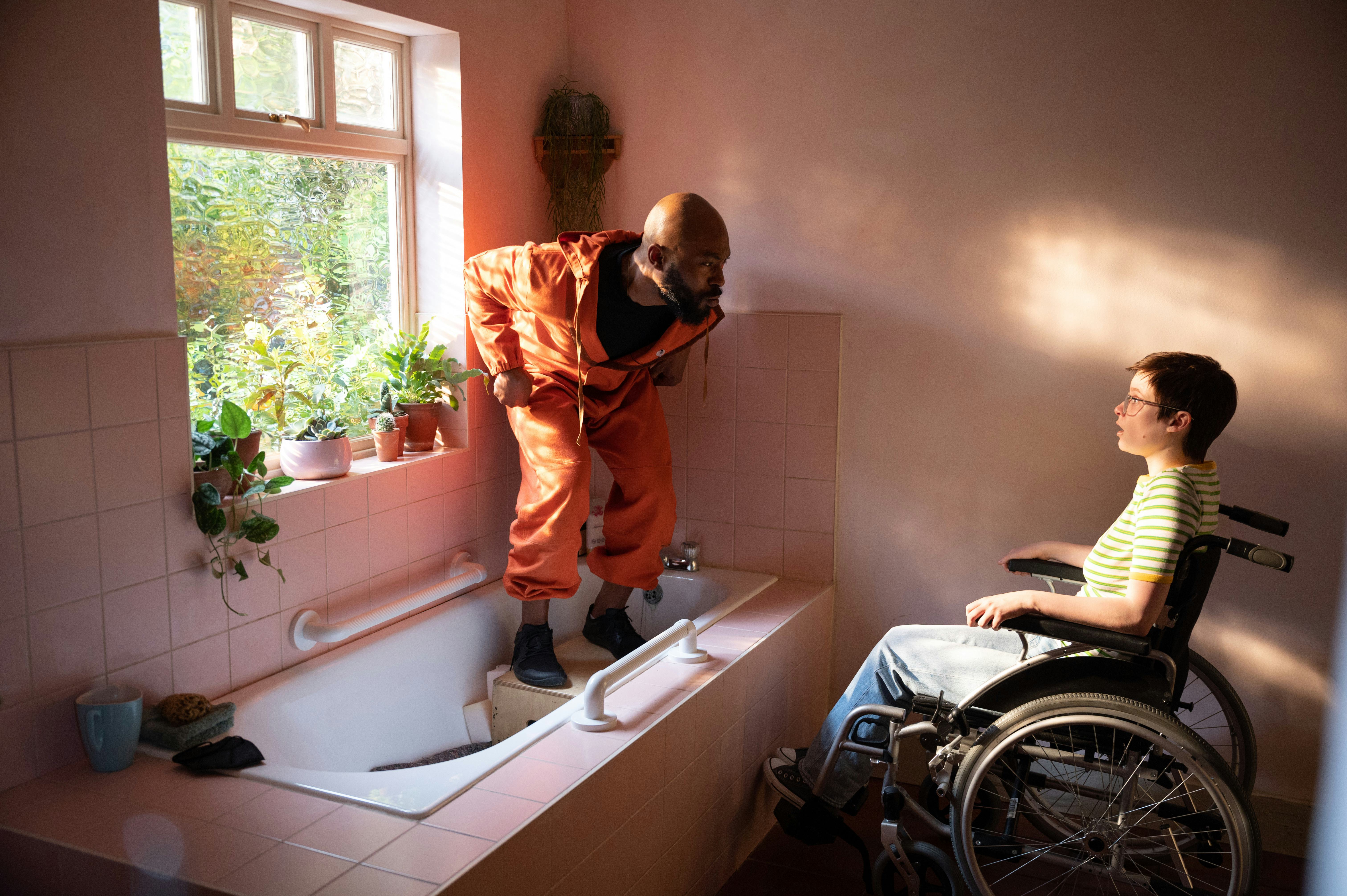 Arinzé Kene in orange tracksuit standing in bathtub talking to Lola Petticrew, who plays Tuesday, in&hellip;