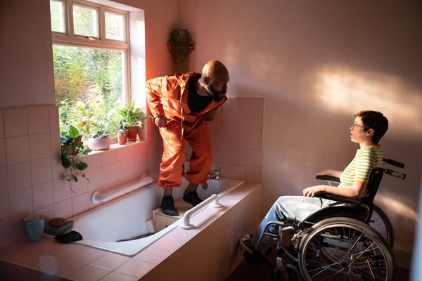 Arinzé Kene in orange tracksuit standing in bathtub talking to Lola Petticrew, who plays Tuesday, in…