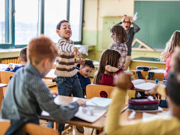 Children cause chaos in a classroom as a teacher looks on.