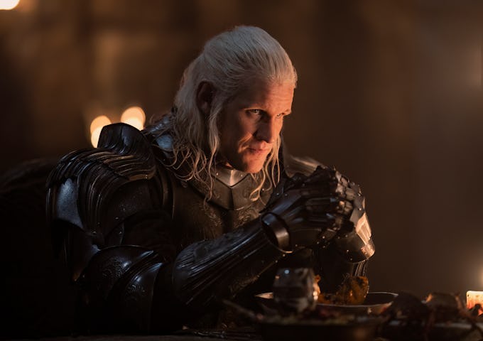 A white-haired man in dark medieval armor sits at a dimly lit table, looking intently while clasping his hands.