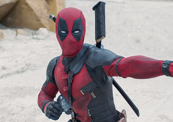 Deadpool in red and black costume pointing while holding a sword, set against a sandy background.