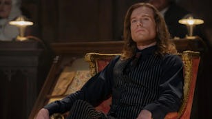 Man with long brown hair seated regally in a striped suit on a golden throne, exuding confidence in an opulent room.