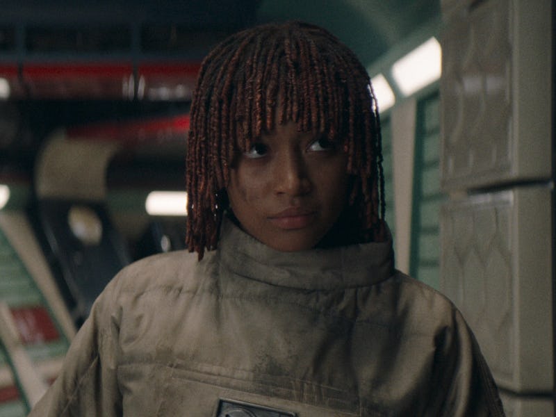A young woman with braided hair, wearing a beige futuristic uniform, stands inside a dimly lit space...