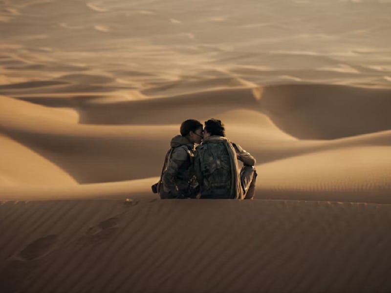 Two people sitting close together in a desert, surrounded by sand dunes, with backpacks, sharing a q...