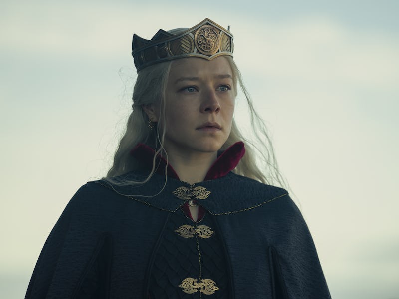 A solemn young woman wearing a golden crown and a dark blue robe stands against a cloudy sky.