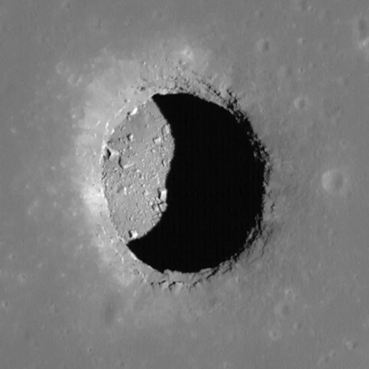 A crescent-shaped shadow covers a third of a hole on the Moon's surface. Big chunks of rock lie insi...