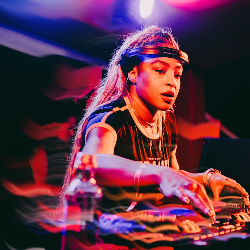 A focused DJ mixes tracks on stage under colorful lights, wearing a headband and earrings.