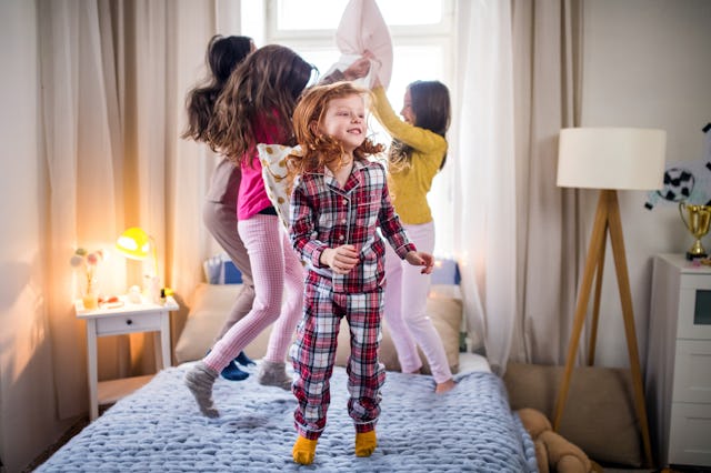 Three young girls joyfully jumping on a bed in a bright, cozy bedroom, one wearing a plaid jumpsuit ...