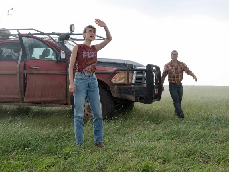 A man and a woman look off into the distance. Their tech-heavy car is parked behind them. The sky is...