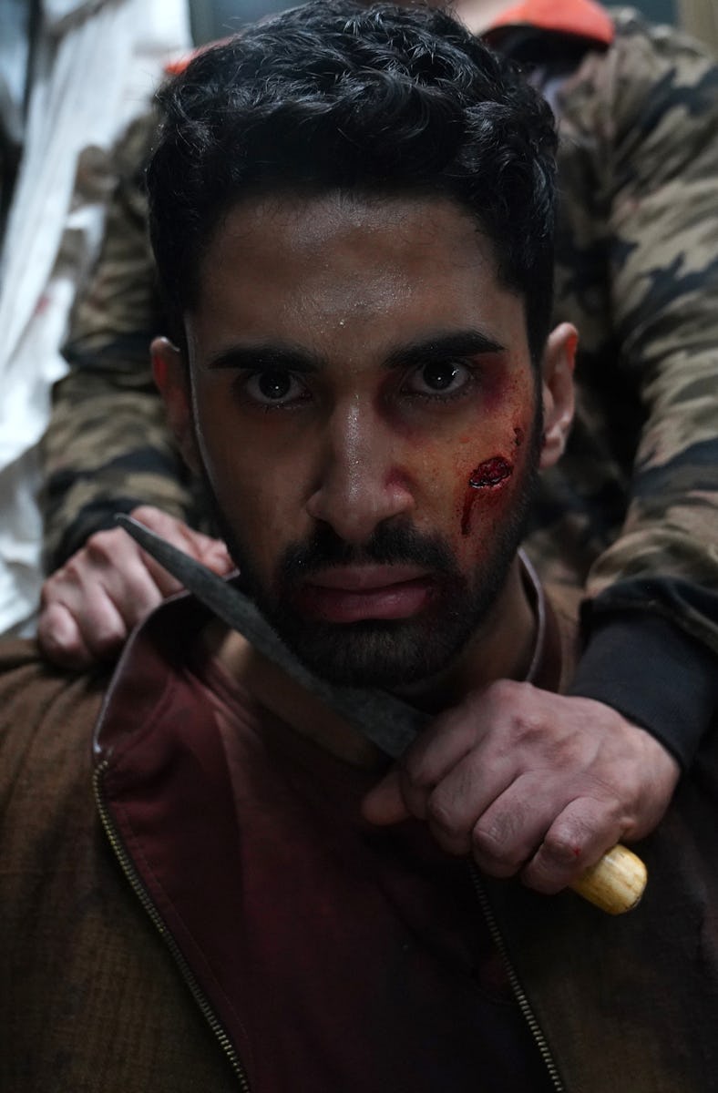 A man with a bloodied face holds a knife, looking intense, seated in a dimly lit, chaotic environmen...
