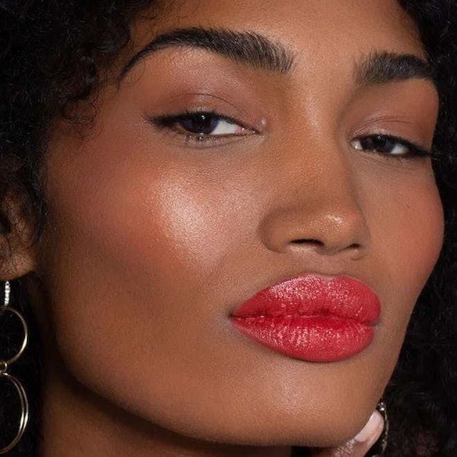 Close-up of a woman with curly hair, wearing Ilia Color Block Lipstick in Grenadine, large earrings, and a thoughtful expression.