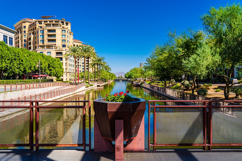 The image shows a view from a bridge over the Arizona Canal in Scottsdale showing some of the buildi...