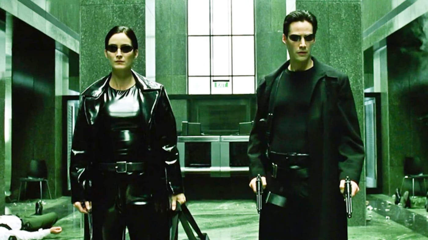 A man and a woman dressed in black leather outfits stand in a damaged, green-tinted room, displaying a defiant look.