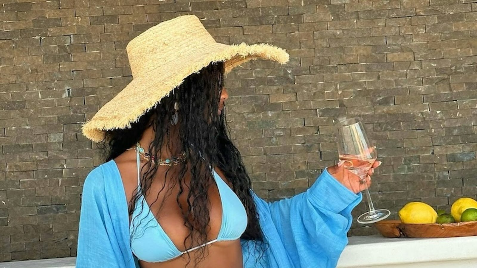 stands against brick background wearing blue string bikini with matching cover up and sarong with wide-brim straw hat, wine glass in hand