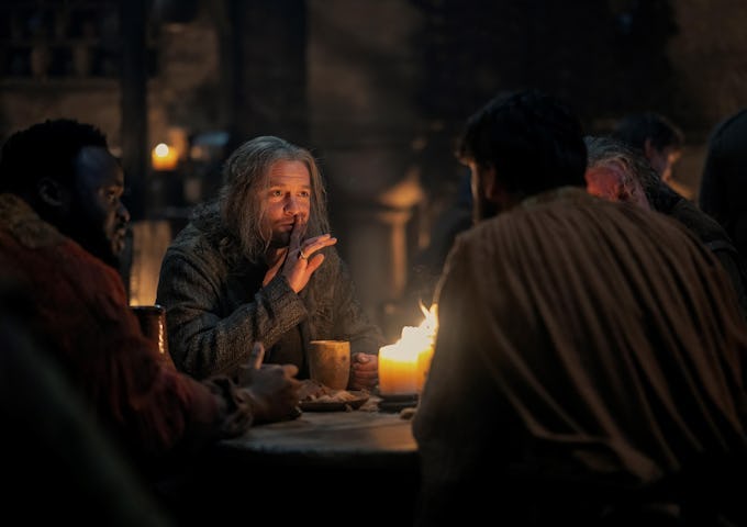 An elderly man with a long gray beard speaks calmly at a dimly lit, medieval-style dinner table surrounded by other people.