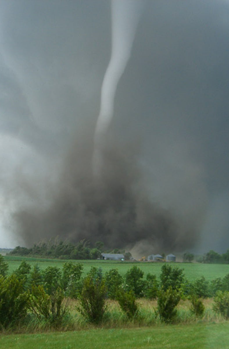 A tornado funnel sweeps up dirt and debris. The dark cloud towers behind farm buildings in the dista...