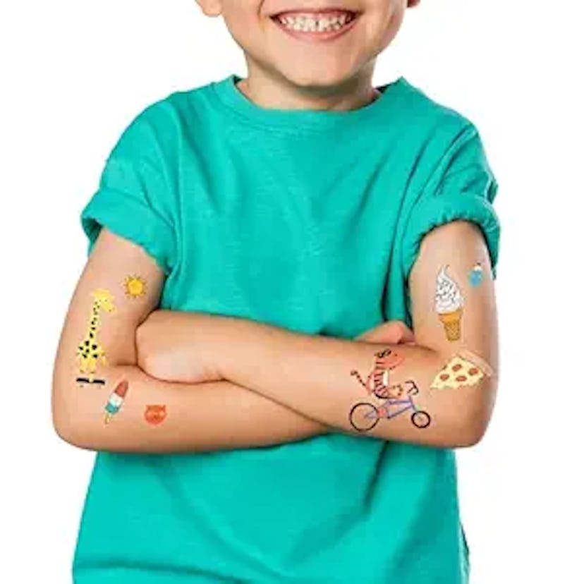 Young boy in a teal shirt smiling with arms crossed, showing off colorful temporary tattoos on his a...