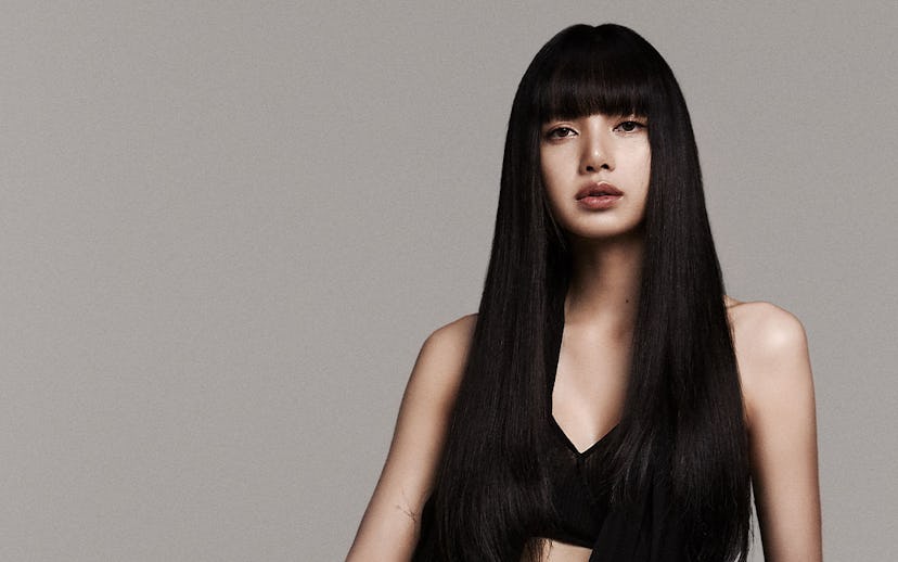 Woman in black jumpsuit with long straight hair sits on a trunk, posing elegantly against a grey bac...