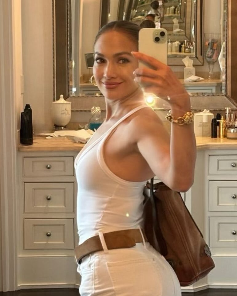 Jennifer Lopez taking a mirror selfie in an all white outfit with brown and gold accessories.