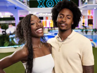 A cheerful couple posing with smiles in a colorful room with neon lighting and "XOXO" sign in the ba...