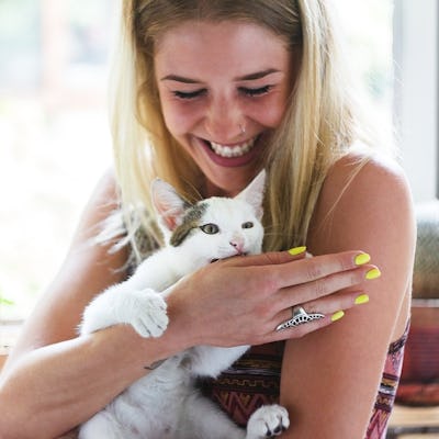 A joyful woman holding a white and gray cat near a window with plants in the background.