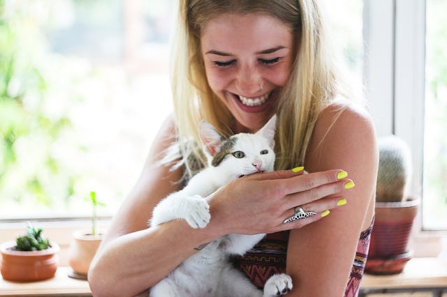 A joyful woman holding a white and gray cat near a window with plants in the background.