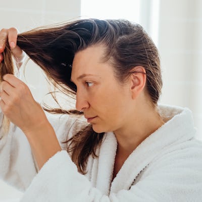 A woman in a white bathrobe inspects her hair in a brightly lit bathroom.