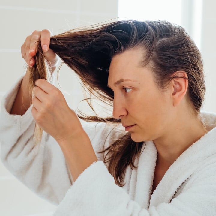 A woman in a white bathrobe inspects her hair in a brightly lit bathroom.