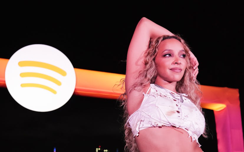 A performer with curly hair, dressed in a white outfit, poses on stage with a city skyline backgroun...