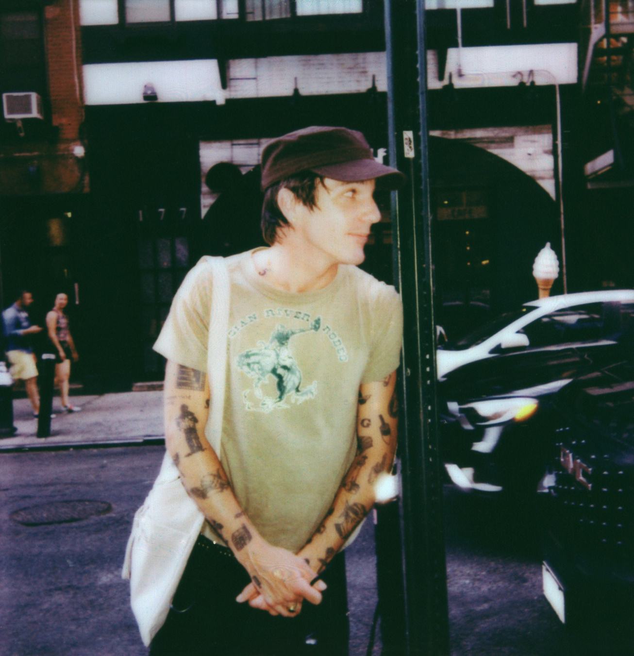 Man with tattoos, wearing a cap and a green t-shirt, leaning against a post on a busy street.