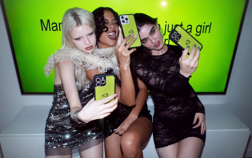 Three women in stylish outfits taking selfies with their phones in front of a neon-lit sign reading ...