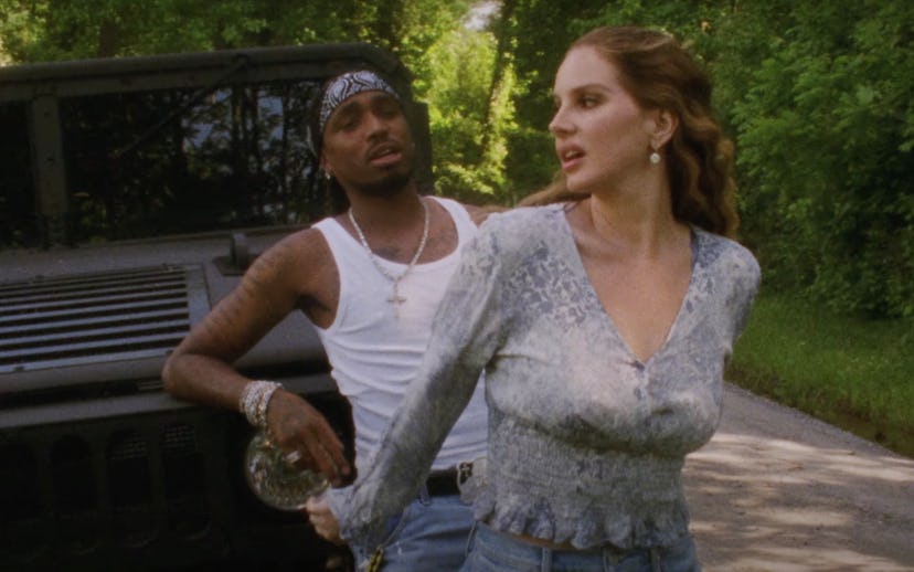 A man and a woman walking near a military vehicle, the man in a white tank top and bandana, and the ...