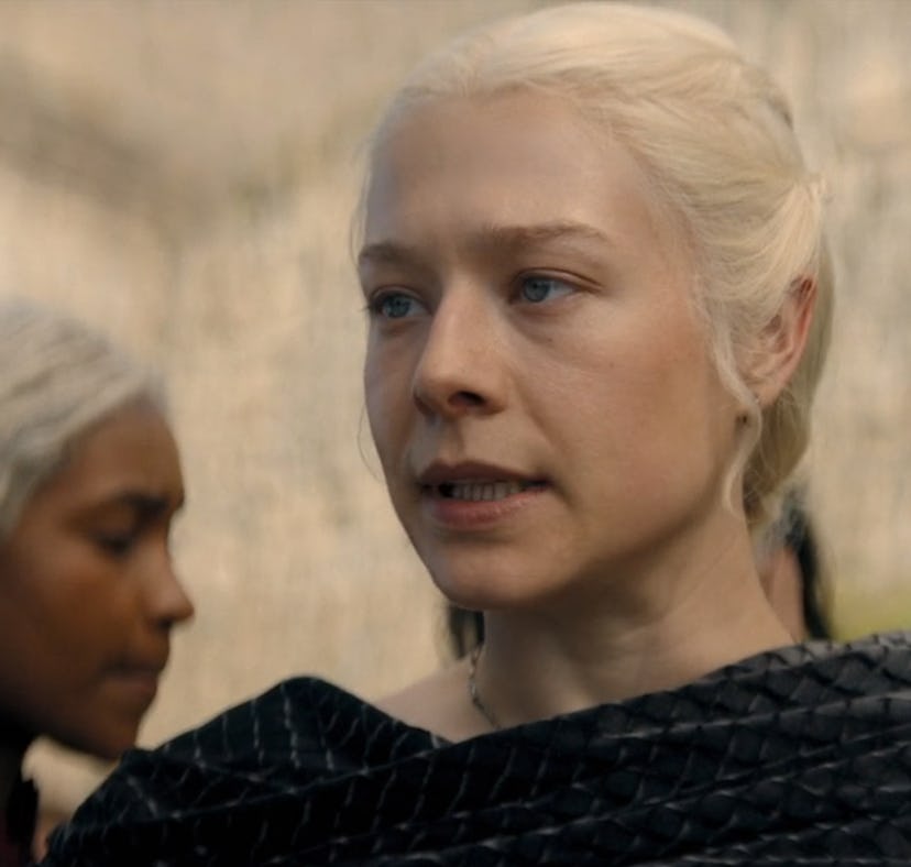 Blond woman with an expression of resolve, wearing black clothing, stands in focus, with another wom...