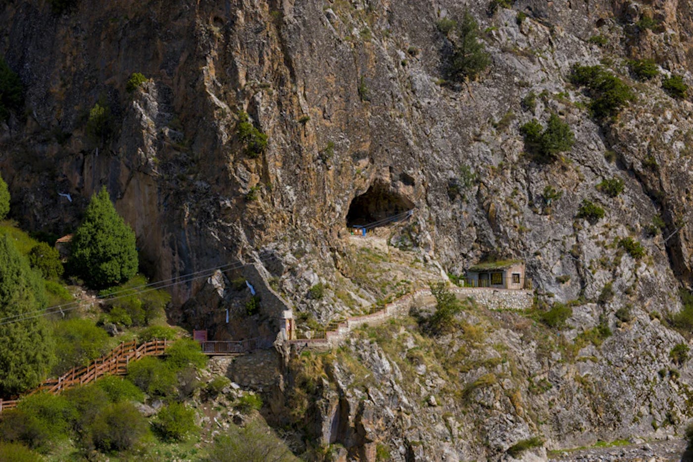 A mountainous landscape with a rugged cliff face featuring a small cave, a wooden pathway, and a tiny stone building surrounded by greenery.
