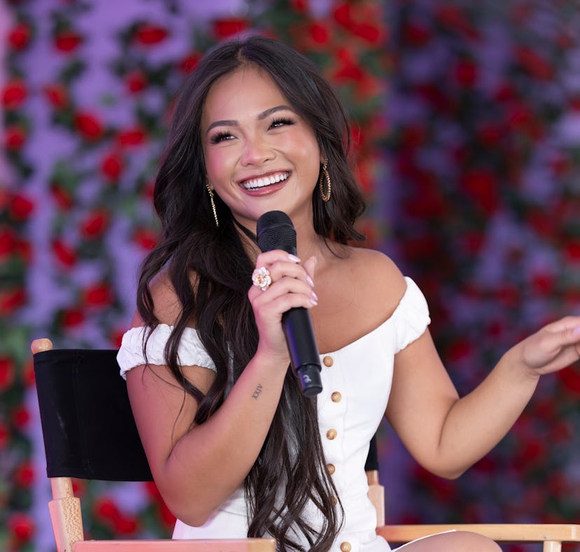 A joyful woman speaking into a microphone, sitting in front of a rose-covered background, wearing a ...