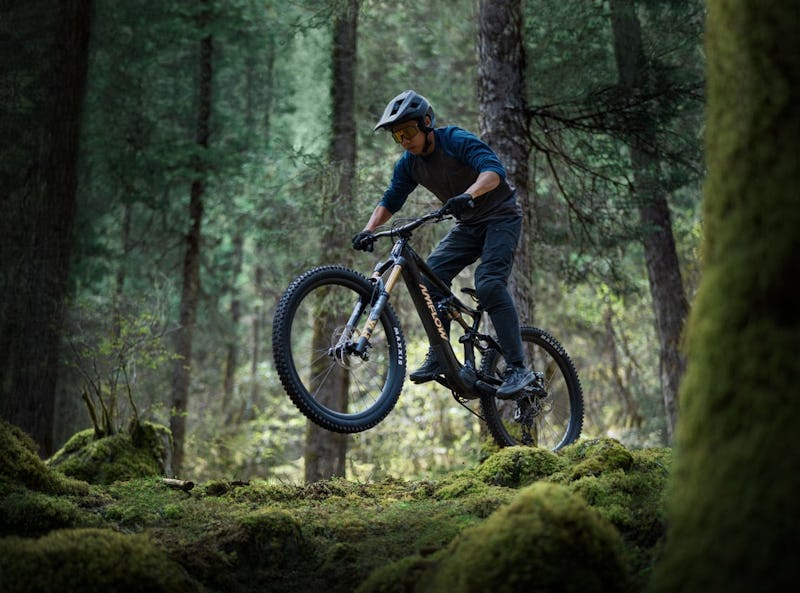A mountain biker performs a jump between moss-covered trees in a lush, green forest.