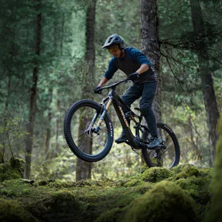 A mountain biker performs a jump between moss-covered trees in a lush, green forest.