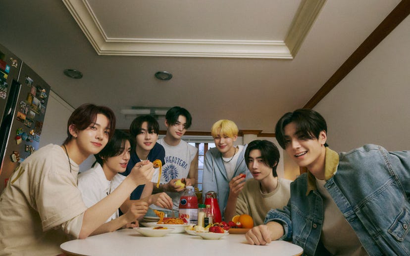 Seven young men smiling around a table filled with food, taking a selfie in a cozy kitchen setting.