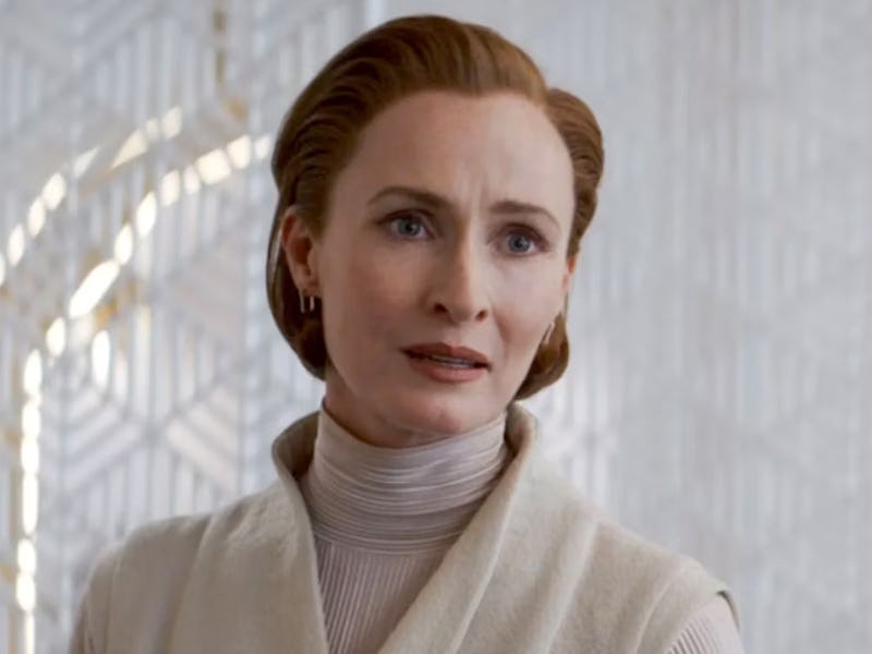 A woman with red hair styled back, wearing a cream turtleneck, looks concerned in a room with intric...