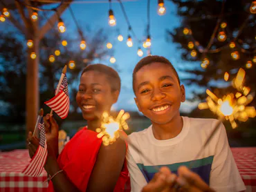 Two joyful teenagers at an evening outdoor party with sparklers and string lights, one holding an Am...