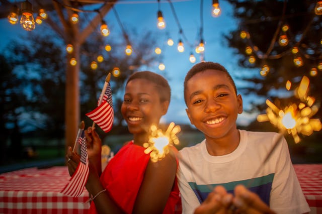 Two joyful teenagers at an evening outdoor party with sparklers and string lights, one holding an Am...